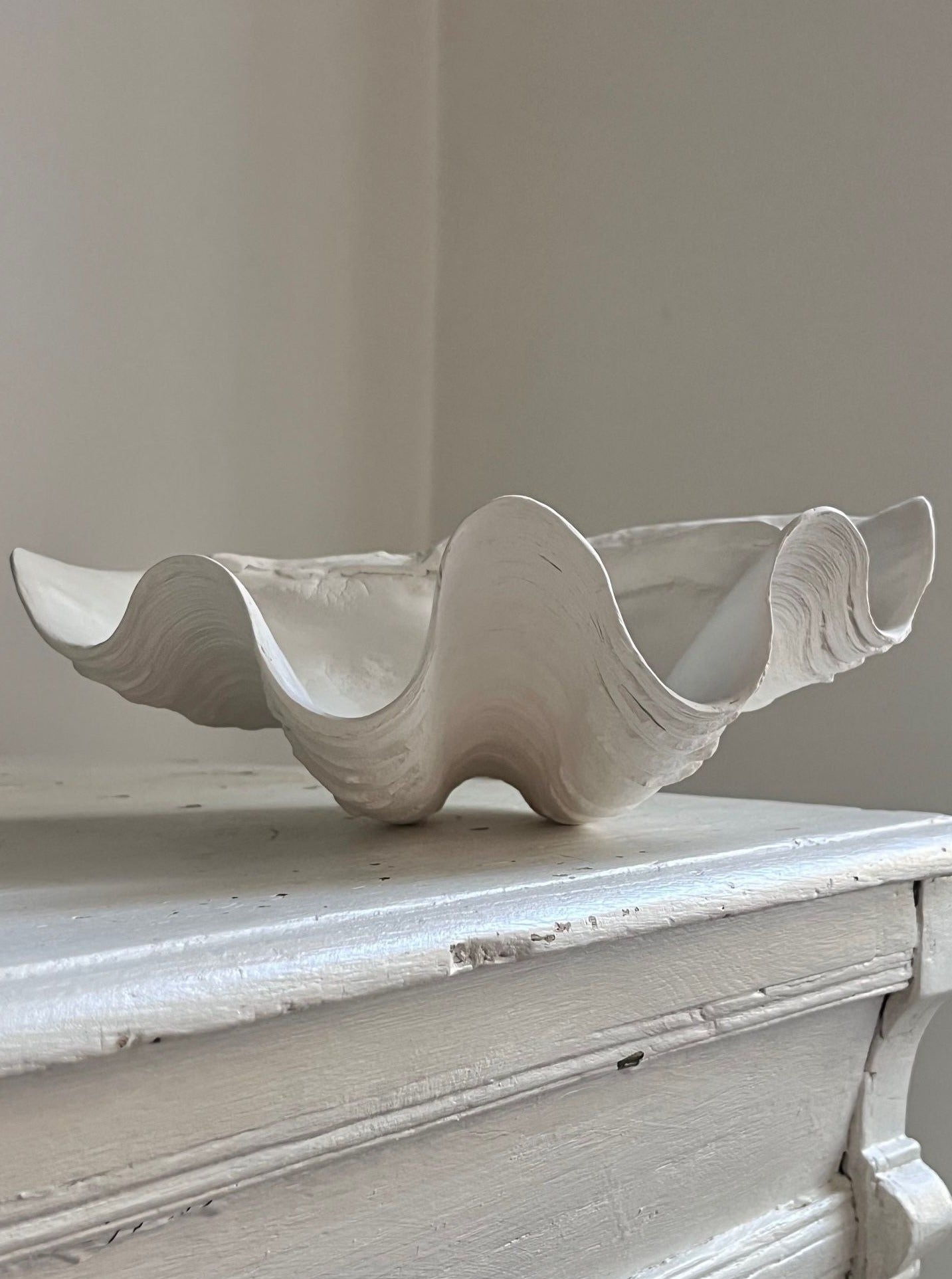 A wavy, sculptural ceramic Médecine clam shell bowl with flowing edges, displayed on a faded white wooden table against a neutral wall background.