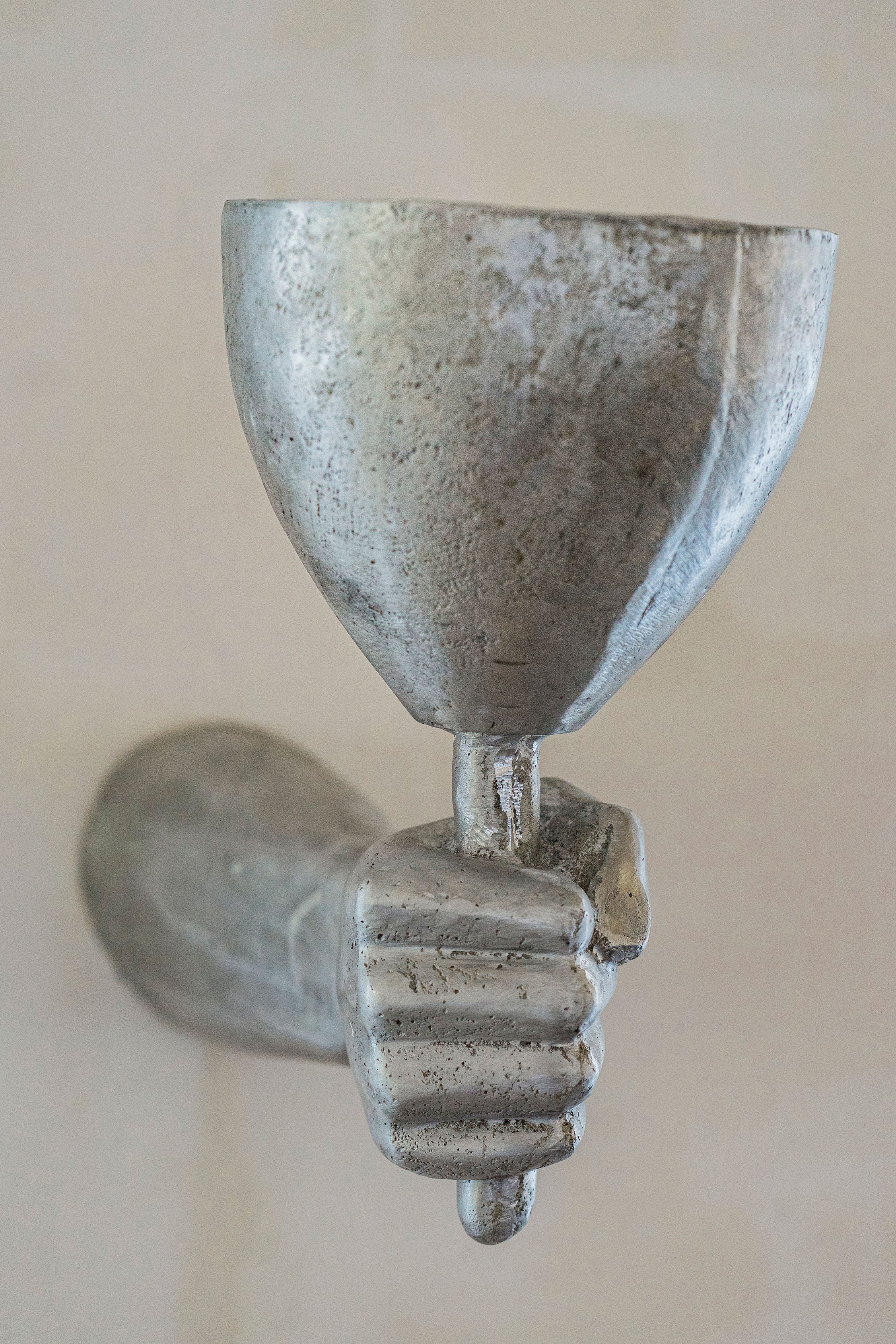 A close-up photo of a weathered sculpture of a hand gripping a chalice, cast in high-quality aluminum, mounted on a wall, with a focus on the textured details of the metal - Rooms Studio's Dracula Wall Scone Aluminium.