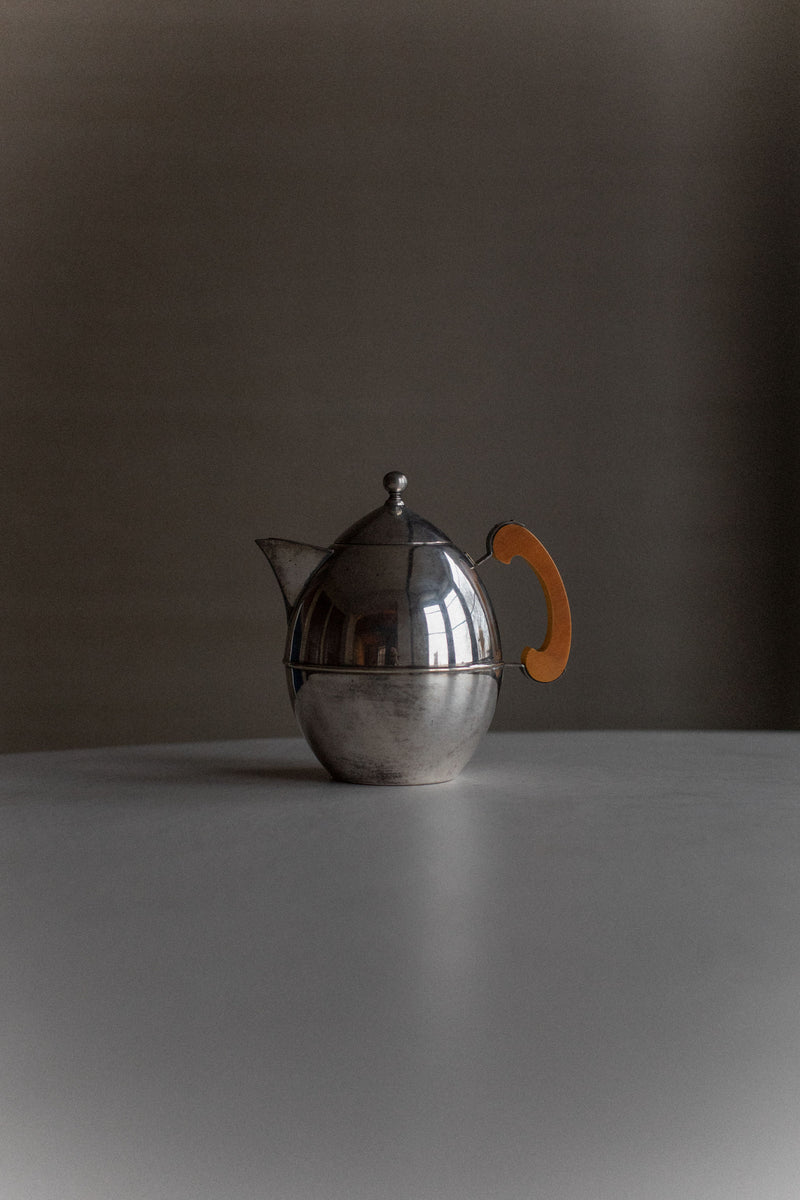 A polished silver-plated Art Deco Teapot with a contrasting wooden handle in a curvaceous shape, standing on a flat surface against a muted grey backdrop by Out For Lunch.