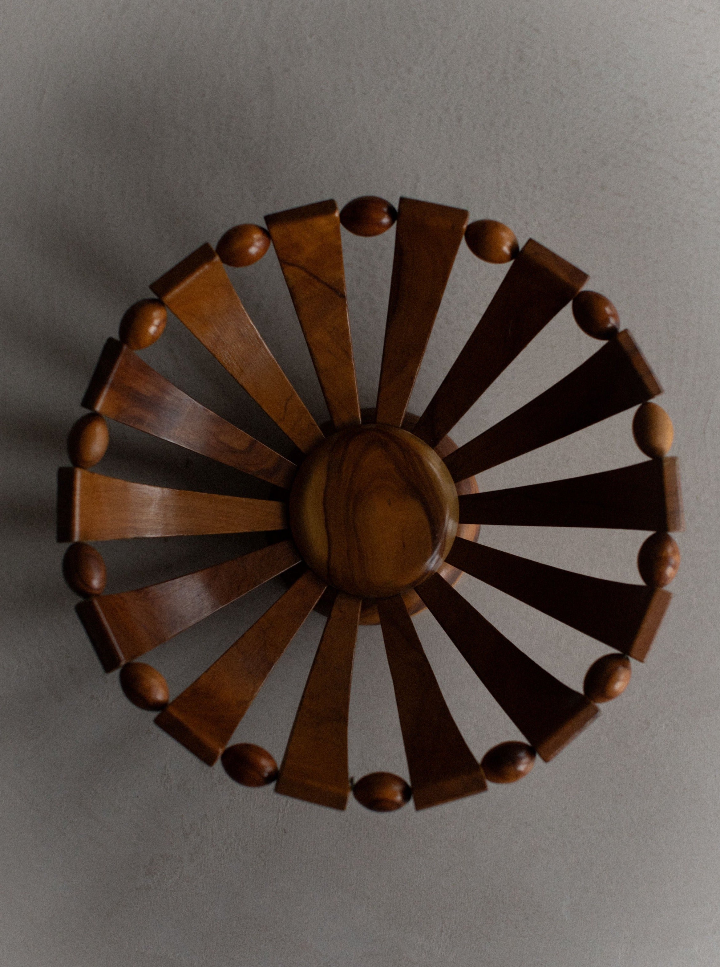 Sentence with replaced product:

An Out For Lunch decorative wooden wall piece shaped like a wheel with alternating light and dark stained wooden slats, showcasing natural grain, and spherical knobs along the edge, mounted on a textured grey wall.