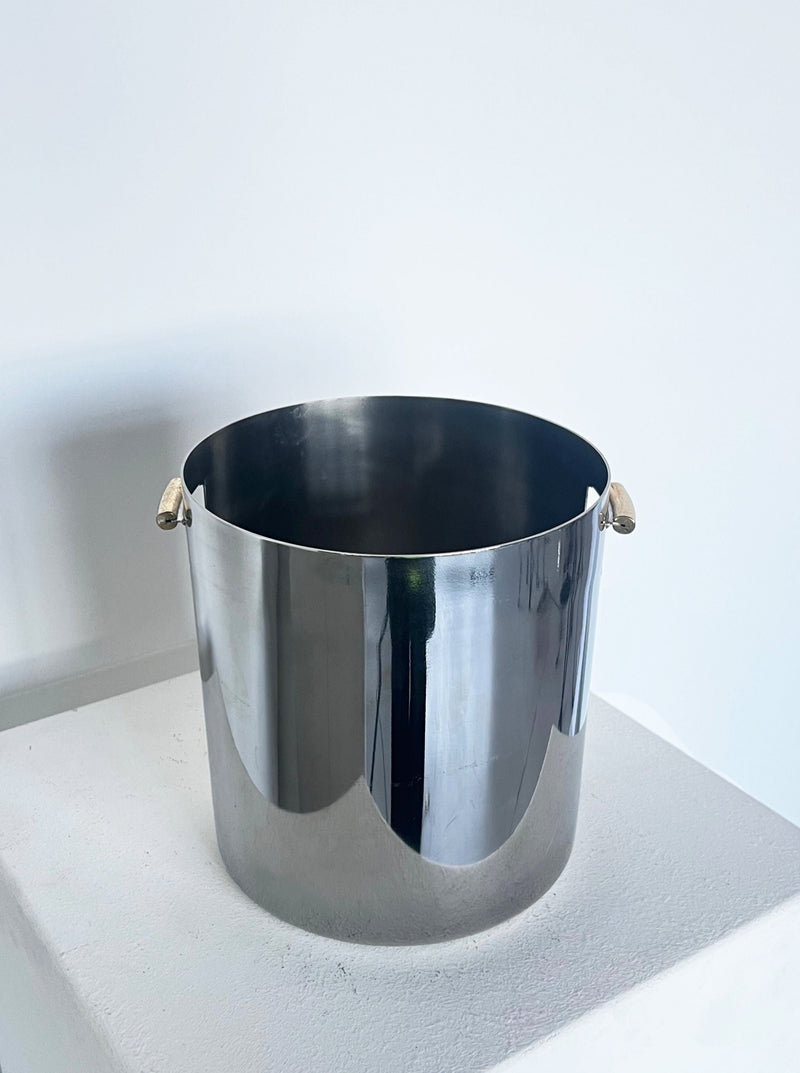 A shiny stainless steel pot with brass handle detail, sitting on a white surface against a white wall. 
would be:
A Brutalist ice bucket with brass detail handle from Porter Studio, sitting on a white surface against a white wall.