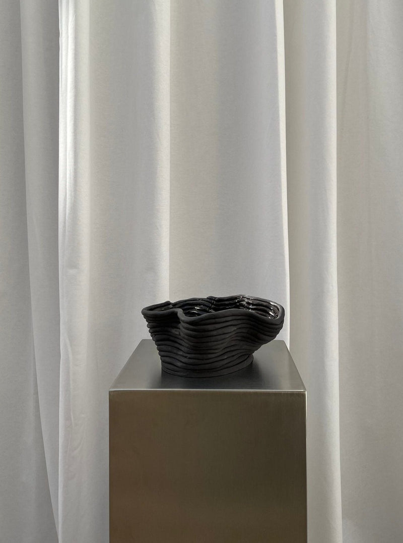 The Wobbly Bowl Black by 2222Studio features wavy edges and a layered design, sitting on a minimalist gray plinth against soft white drapes. Its handcrafted dark texture creates a striking contrast with the smooth backdrop, embodying a minimalist aesthetic.
