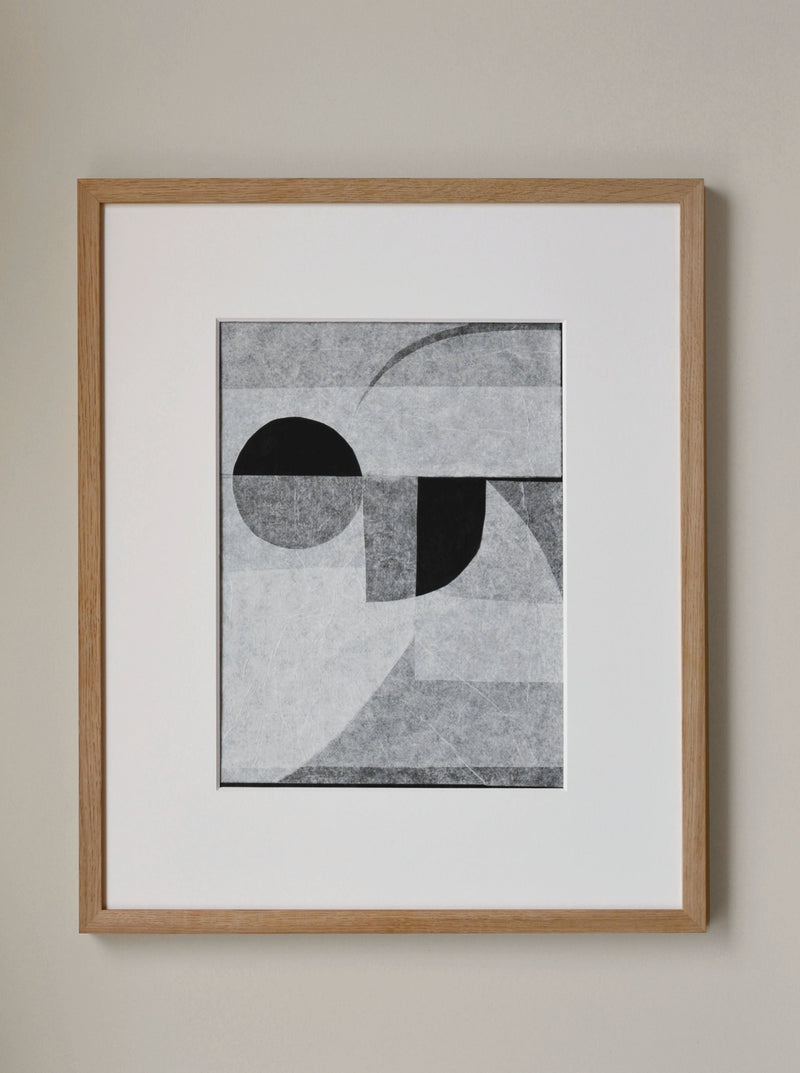 A framed abstract geometric artwork featuring overlapping circles and angular shapes in shades of gray, displayed on a cream-colored wall, created on handmade cotton paper by Julita Elbe's Invisible.