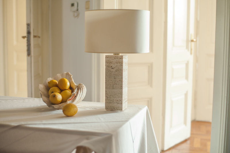 A cozy room with a basket of lemons on a white tablecloth beside a large Brajak Vitberg Capri Travertine table lamp, with an open door in the background. Natural light fills the space, creating a warm atmosphere.