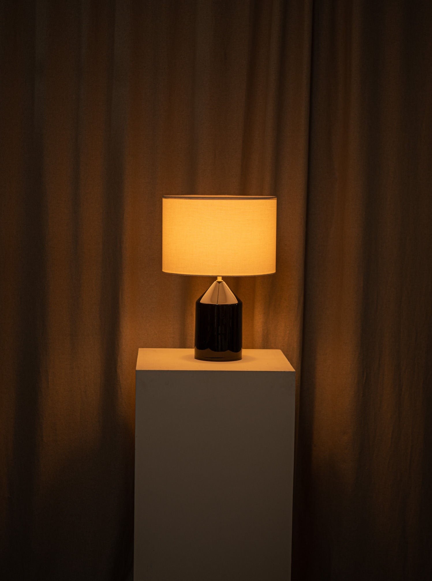 A warm-hued Simone & Marcel Josef Black Ceramic table lamp with a cotton drum lampshade casts a soft glow on a beige pedestal against a draped brown curtain background, creating a cozy and dimly lit atmosphere.