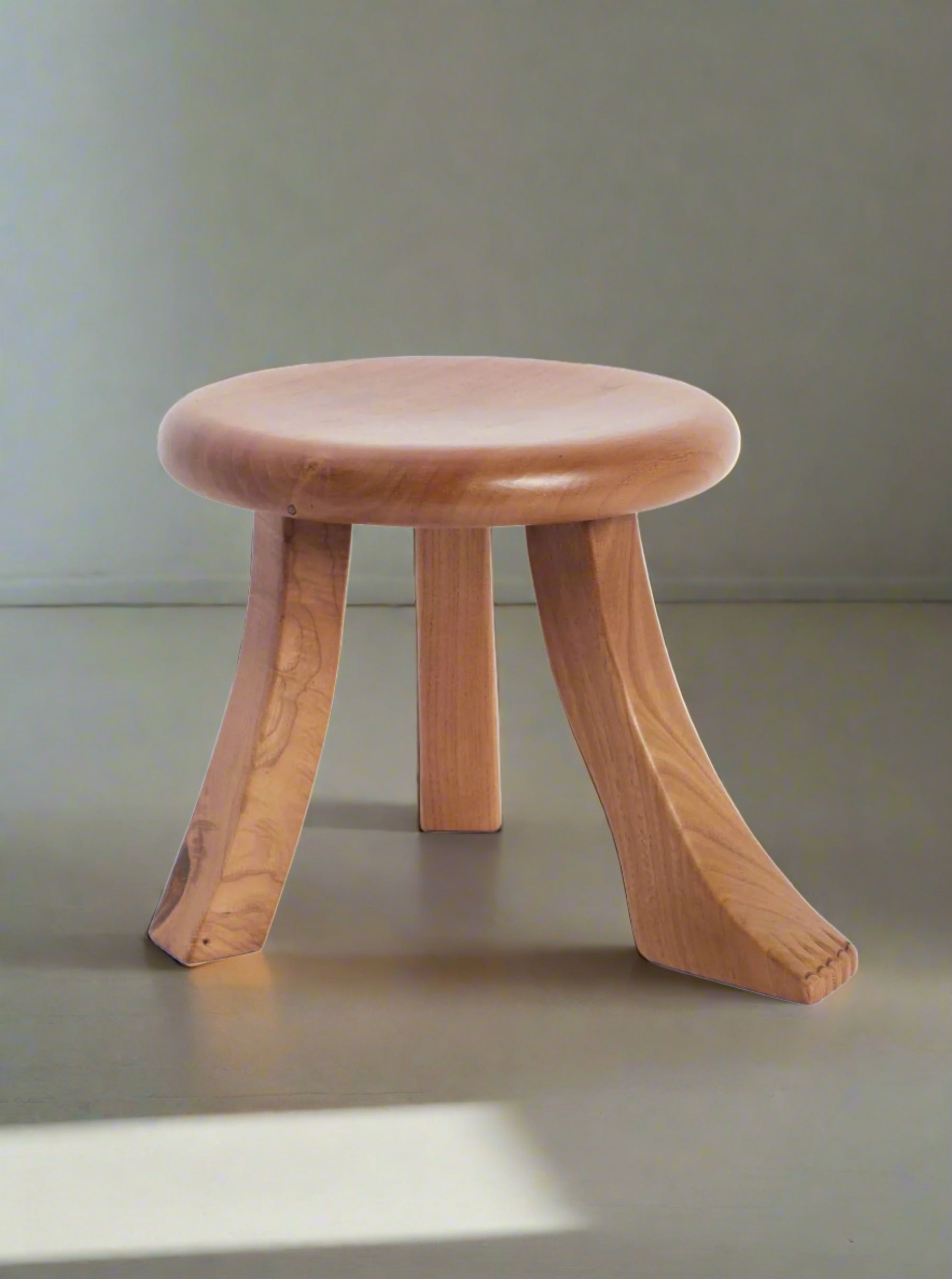 The Foot Stool in Oak by Project 213A, featuring a small, round wooden design with elegantly curved legs and a smooth finish, sits on a light floor and against a light wall. Natural light casts an artful shadow, enhancing its minimalist and collectible furniture appeal.