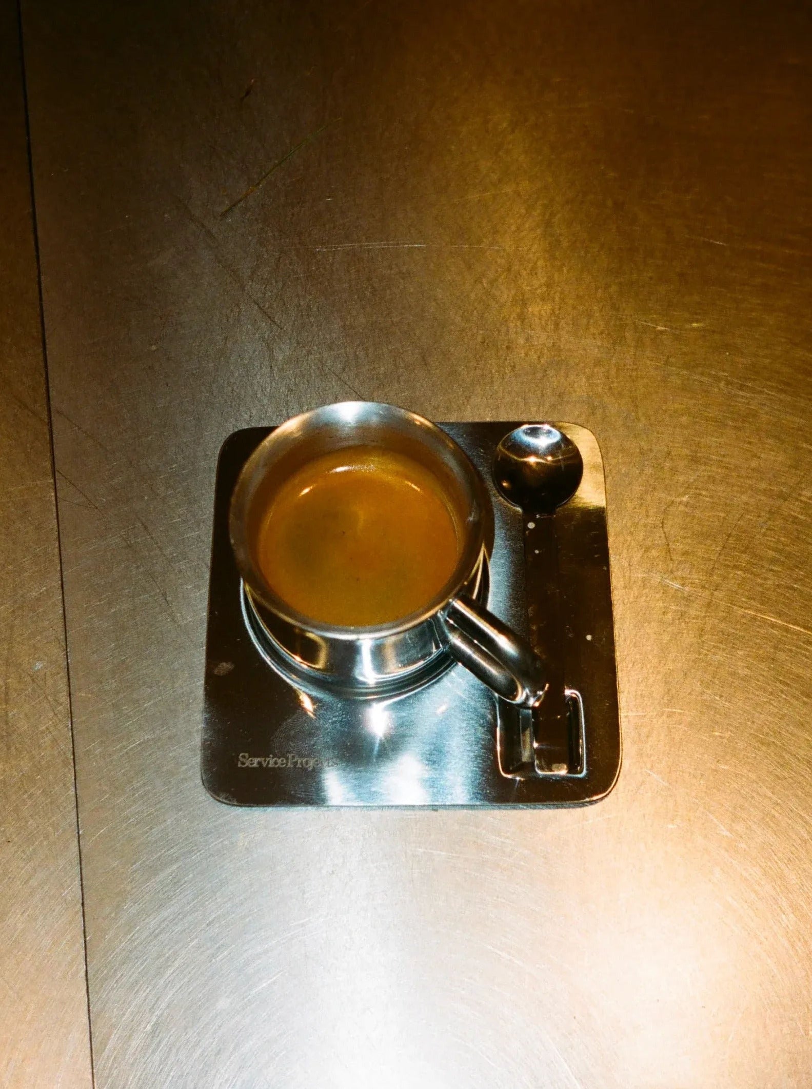 A top-down view of an Arlo Stainless Steel Cup Set filled with coffee, featuring Italian design, resting on a metal saucer with a handle, placed on a brushed metal surface.