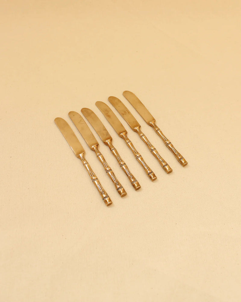 Six brass butter knives with ornate handles arranged in a line on a beige background by Boga Avante Shop.