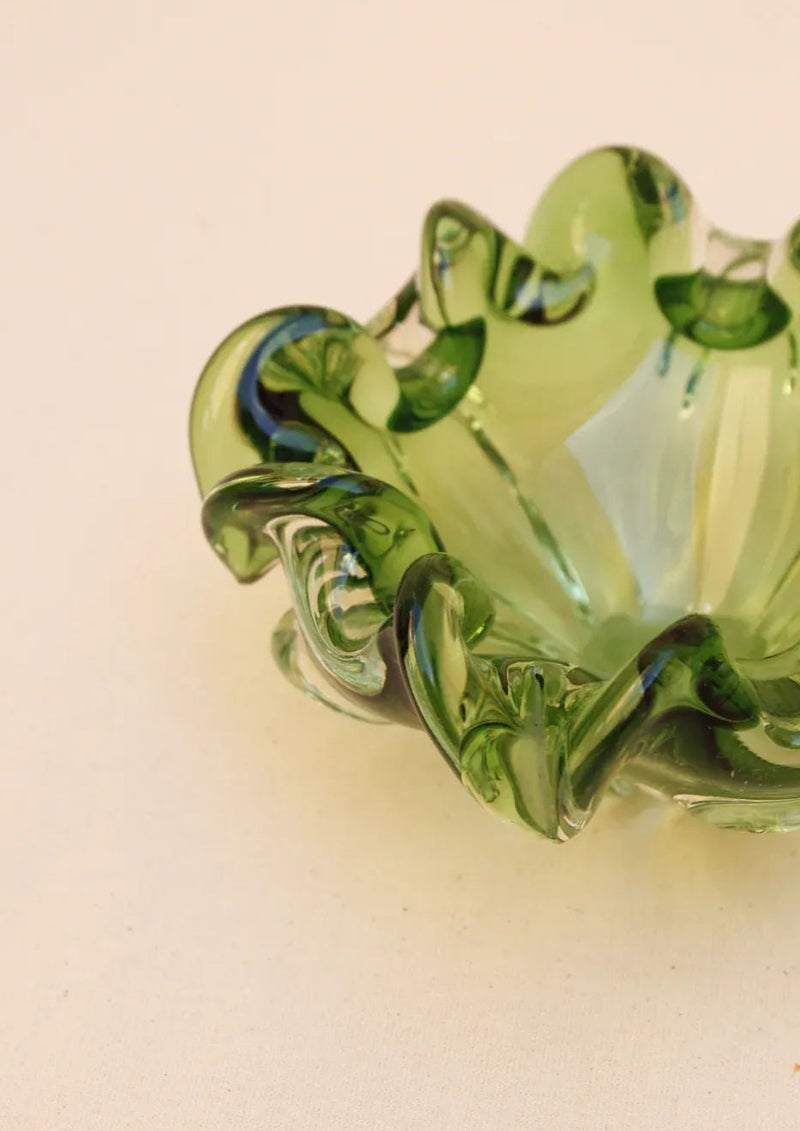 The Murano Glass Bowl from Boga Avante Shop is a handcrafted green bowl with wavy edges and darker green swirls, boasting a translucent design that exudes luxury against a light beige background.