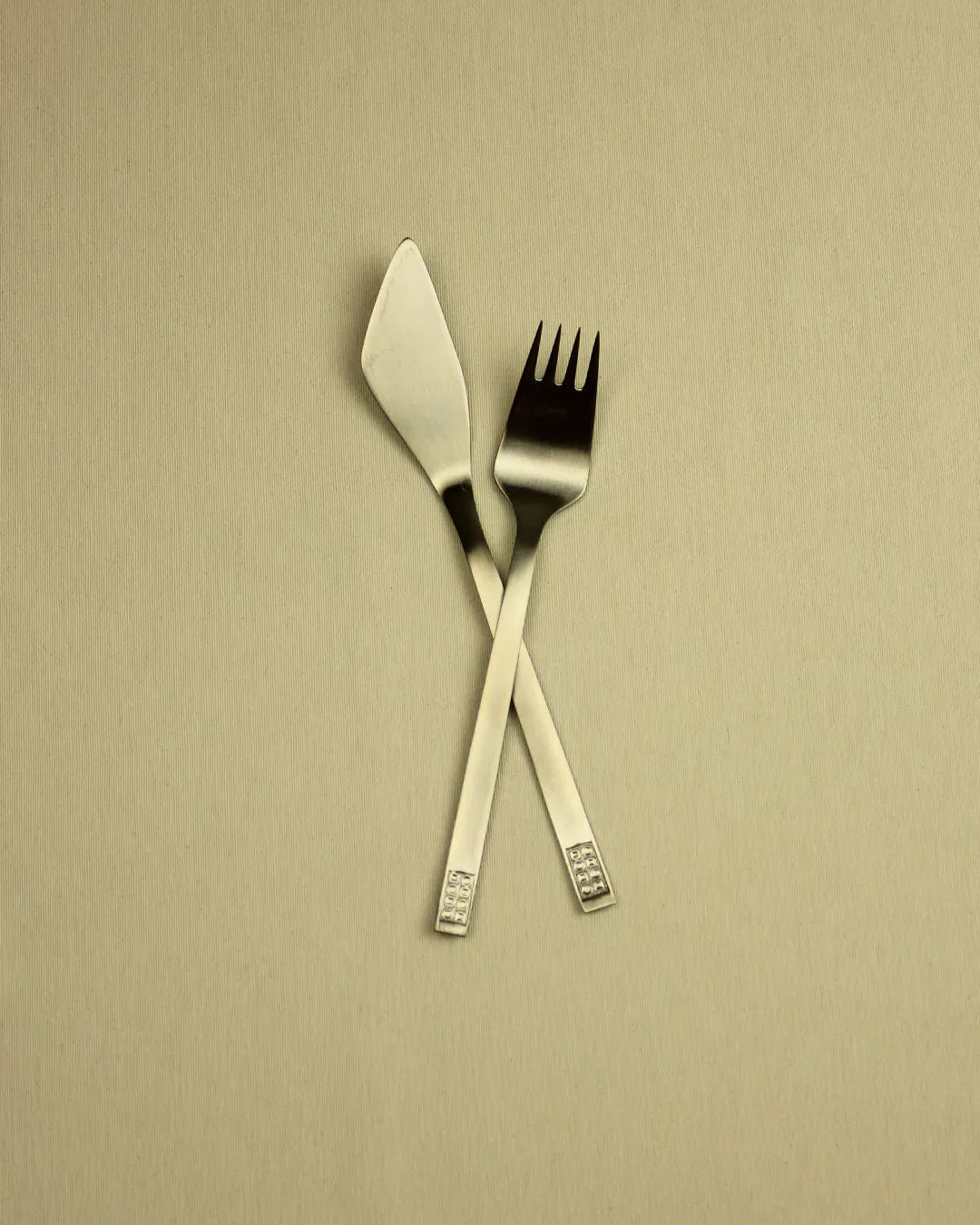 A silver Boga Avante Shop German fish Cutlery Set 70s Style fork and knife crossed over each other on a plain beige background.