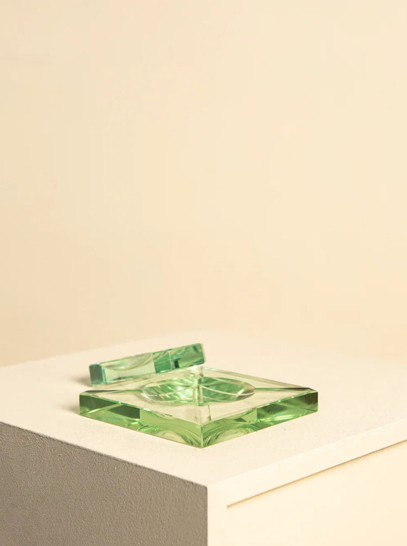 The Glass Desk Set by Fontana Arte 60's from Treaptyque includes a square green glass ashtray with an Italian design, resting on a cream-colored pedestal against a beige background. The ashtray features a small rectangular section for holding a cigarette and embodies an overall simple and elegant aesthetic reminiscent of classic Fontana Arte pieces.