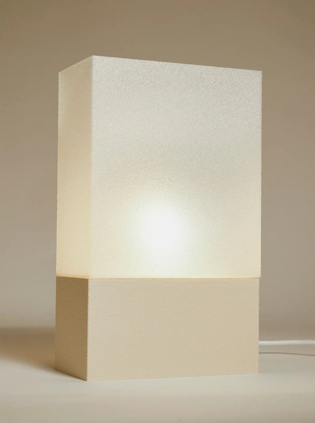 The Lampe i by Argot Studio is a minimalist, rectangular table lamp ideal for luxury interiors. It features a solid, light-colored base with an upper section emitting a soft, warm glow, making it an exquisite piece of collectible furniture that highlights its simple design.