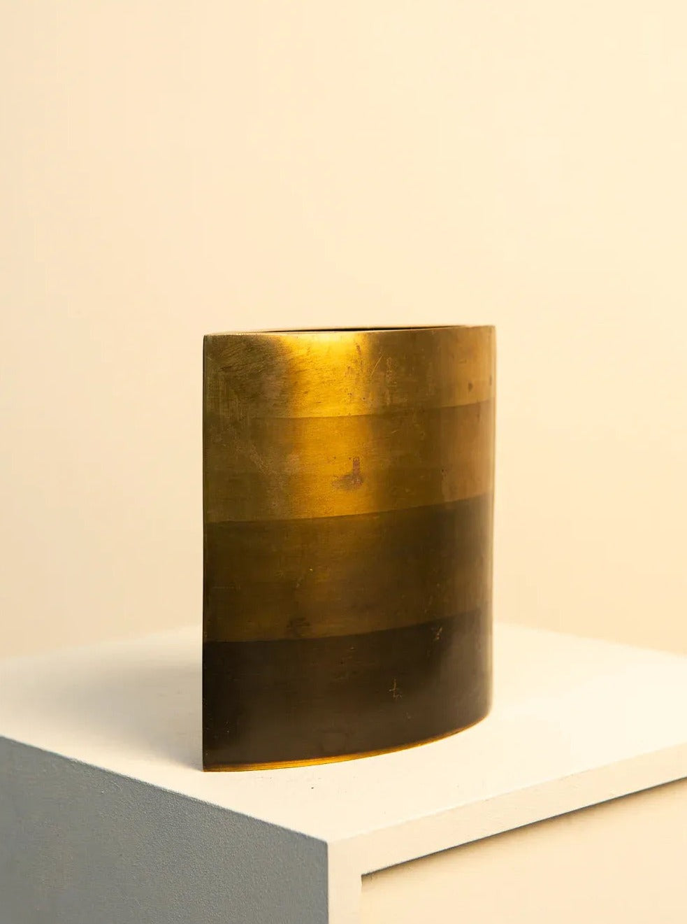 A golden cylindrical Brass and Bronze Vase by Michael Aram 80's on a white ledge against a cream-colored background, illuminated by a warm light, giving it a subtle glow. (Brand Name: Treaptyque)