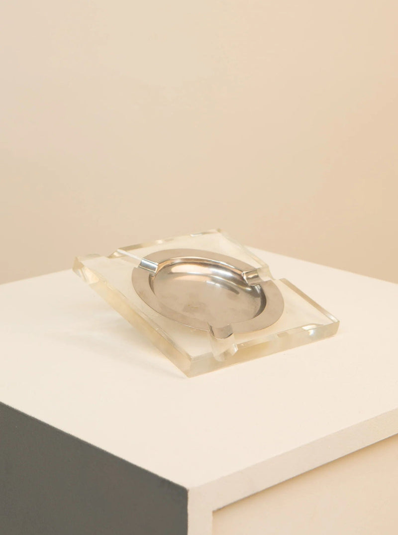 A modern Treaptyque plexiglass and metal 70's ashtray with a metallic inner tray, displayed on a white platform against a beige background.
