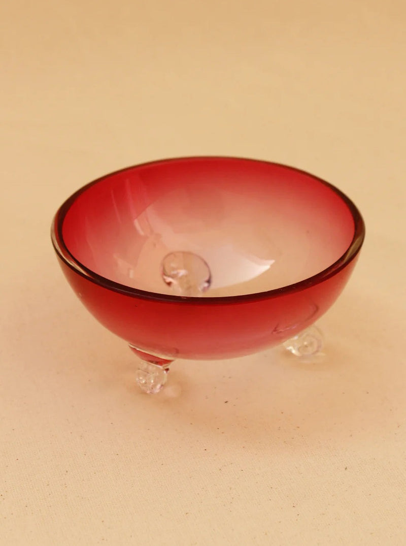The Pink Murano Bowl from Boga Avante Shop is a small, handcrafted glass bowl featuring a round shape and a captivating pink gradient that transitions from dark at the rim to lighter towards the base. Its vintage charm is accentuated by three clear spherical feet, elegantly supported against a plain beige background.
