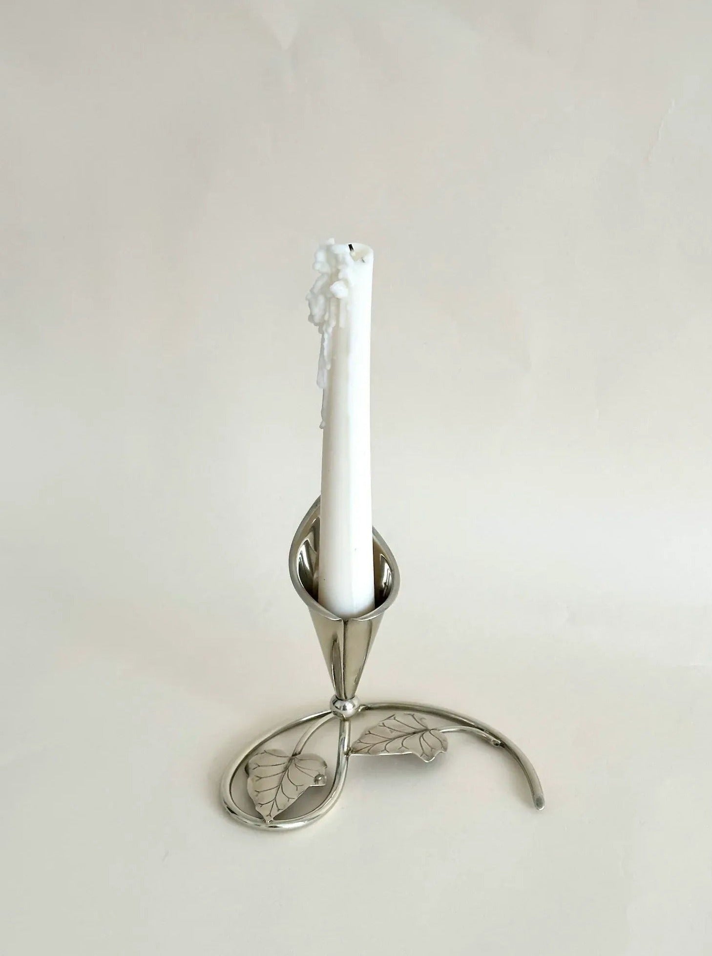 A melting white candle stands in an elegant silver Aesthete Label Cala Lily Candle Holder with botanical-inspired artistry, against a soft beige background.