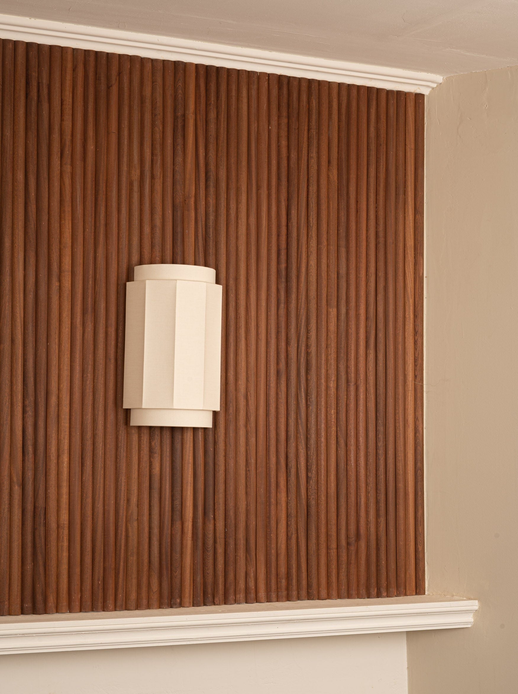 A minimalist beige double wall light from Simone & Marcel mounted on a vertical wood paneling wall painted in varying shades of brown, located in a corner with cream-colored trim.