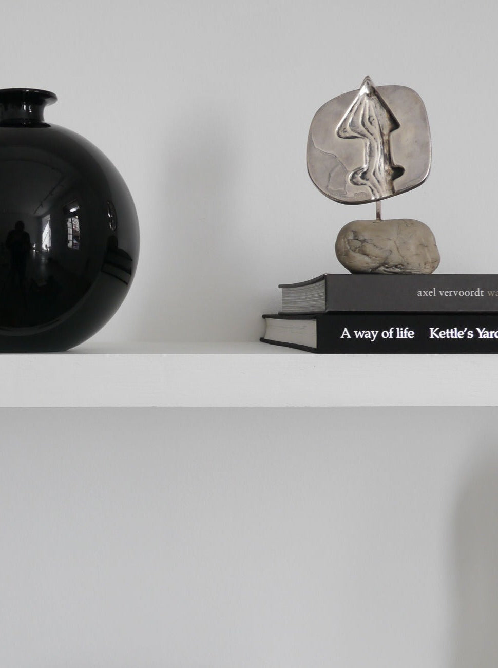 A minimalist shelf displaying a large black vase, two stacked books titled "a way of life" and "De Vecchi Milano," and a small Collection apart Silver Sculpture of a flame-like shape mounted on a marble base