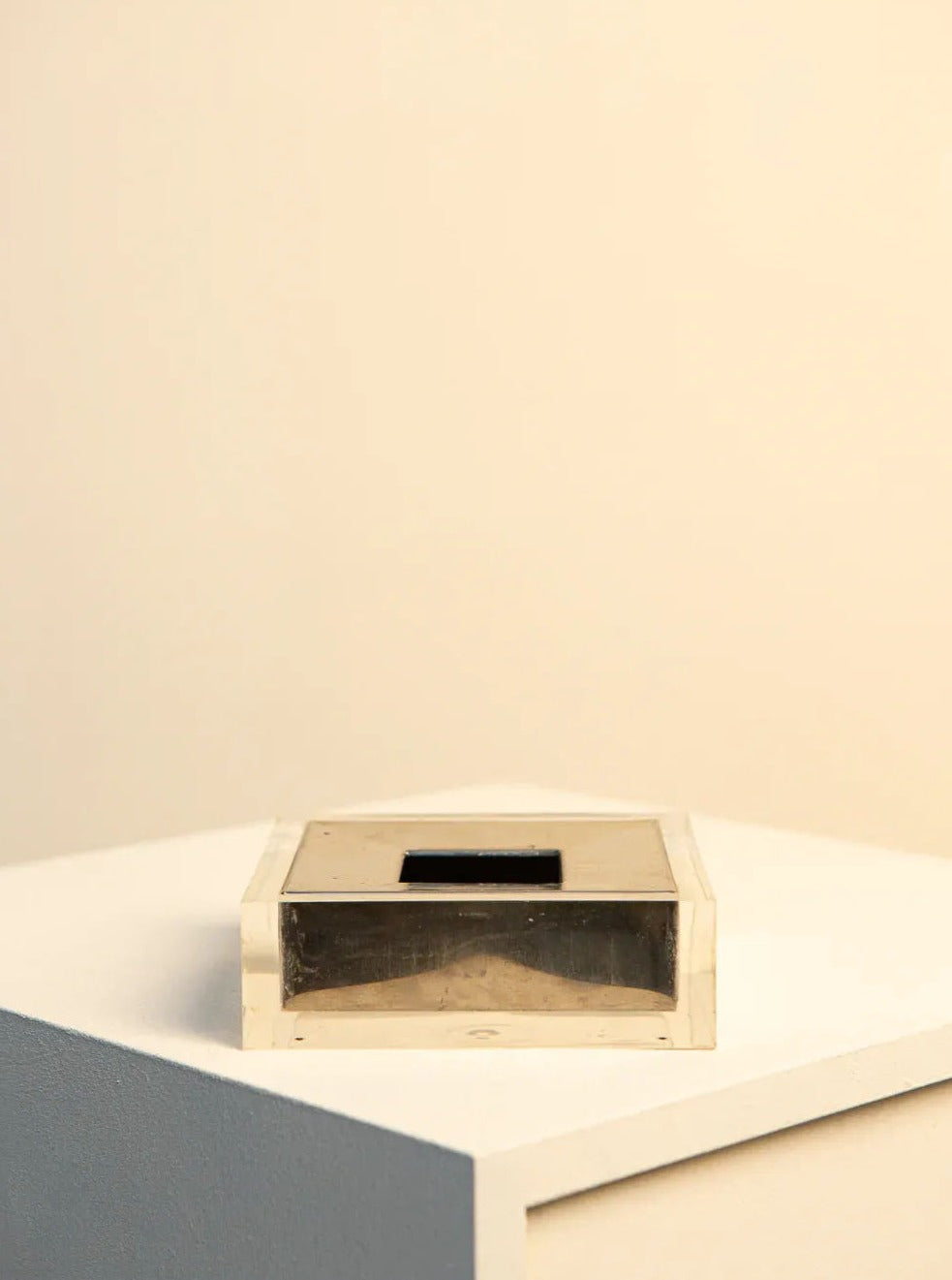 A transparent rectangular container housing a dark object rests on a white geometric surface against a soft beige backdrop. This minimalistic setting, enhanced by Treaptyque's distinctive flair, highlights the simplicity and clarity of Italian design presented in the Metal and Plexiglas Ashtray by Guzzini from the 70s.