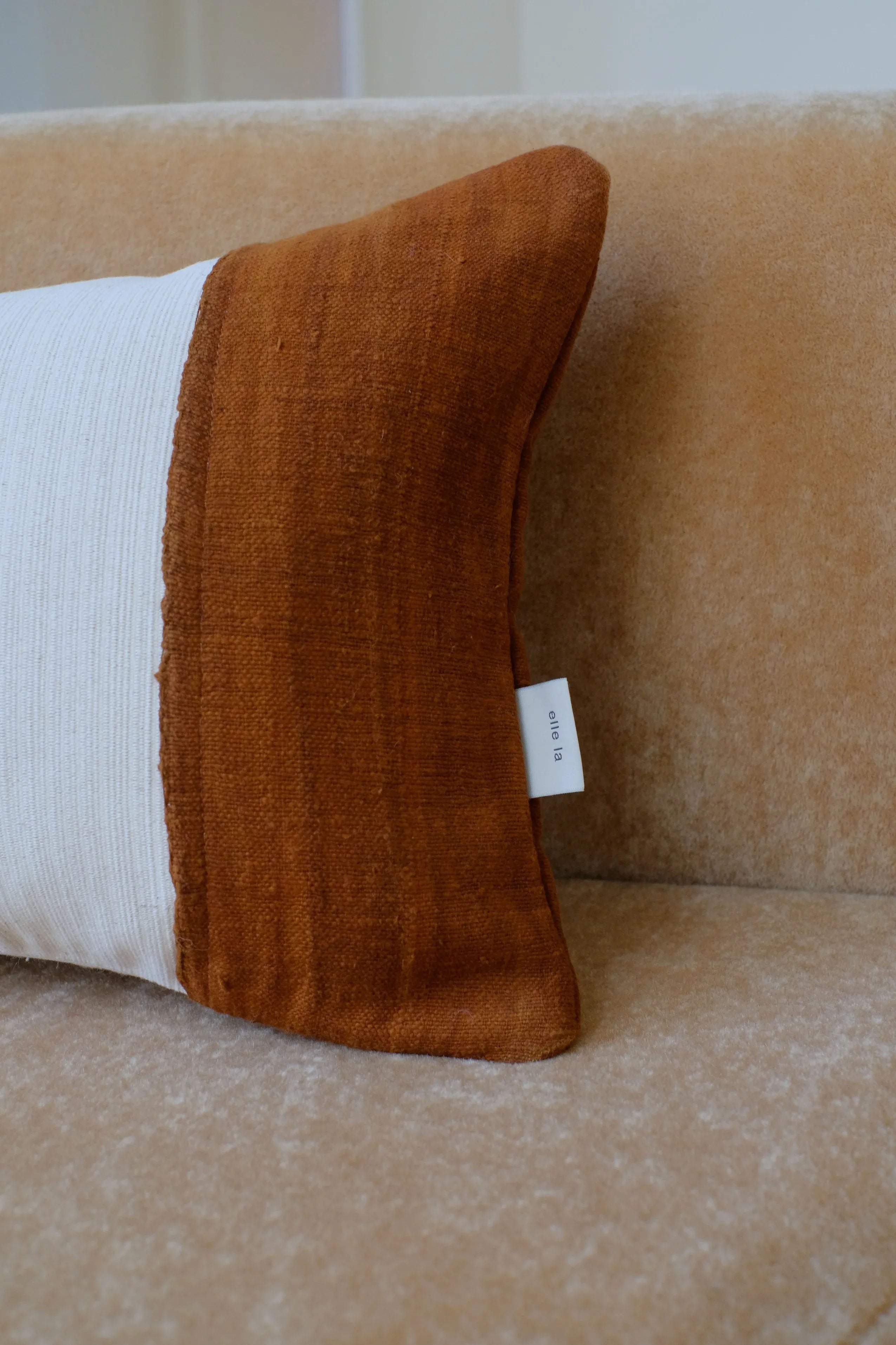 A close-up of an Elle La Luca Longue Cushion with a burnt orange front and a white back against a tan sofa. The handmade-in-Amsterdam cushion has a visible label tag on the side.