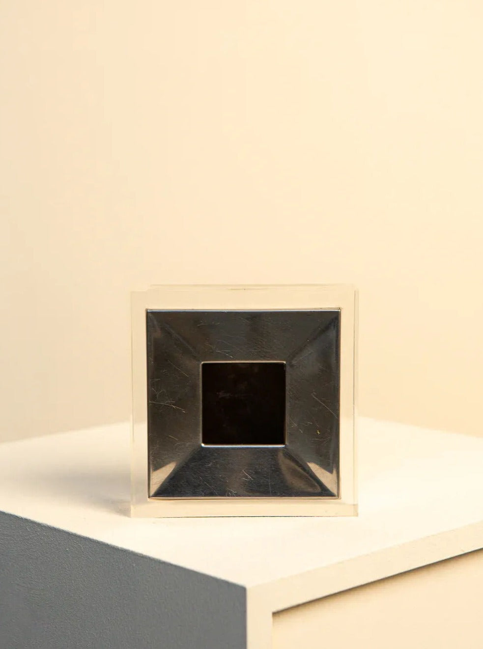 A small, square, metallic object with a reflective surface, identified as the Metal and Plexiglas Ashtray by Guzzini 70's from Treaptyque, sits on a two-toned platform. Reminiscent of Italian design, it features a dark square center and light reflecting off its beveled edges to create a geometric pattern. The background displays a soft gradient of cream to light beige.