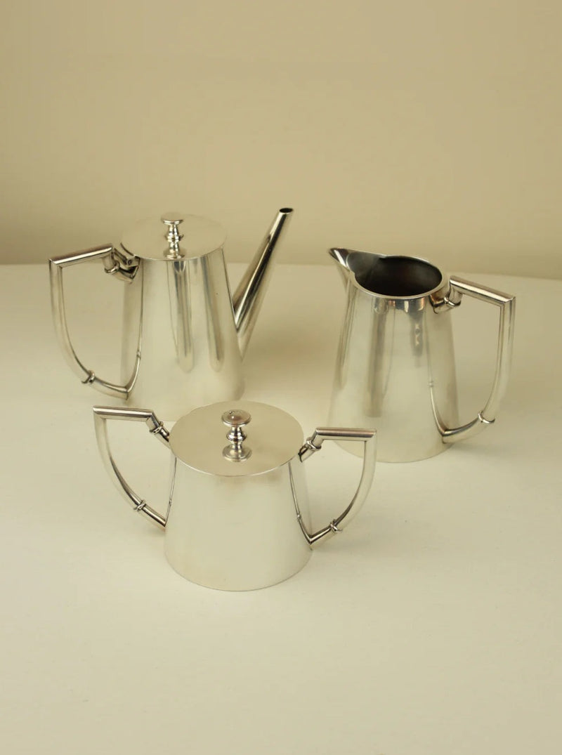 Three Art Deco tea/coffee pots with angular handles and spouts, set against a cream background. The pots vary in size and are part of a vintage coffee service from Boga Avante Shop, reflecting light subtly.