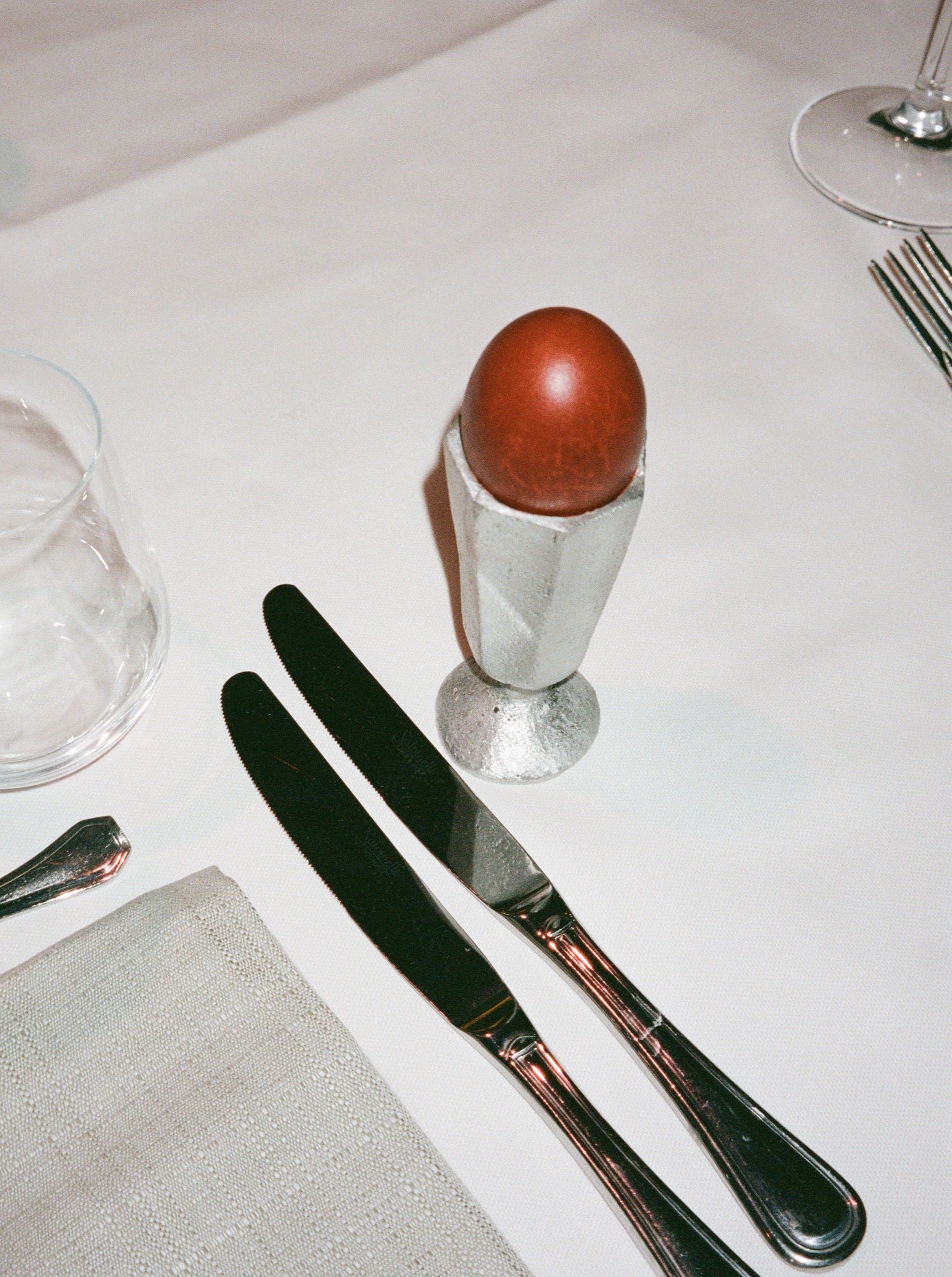 A brown egg in a Rooms Studio high-grade Aluminium Egg Holder placed on a white tablecloth next to a knife and spoon, with a clear water glass partially visible.