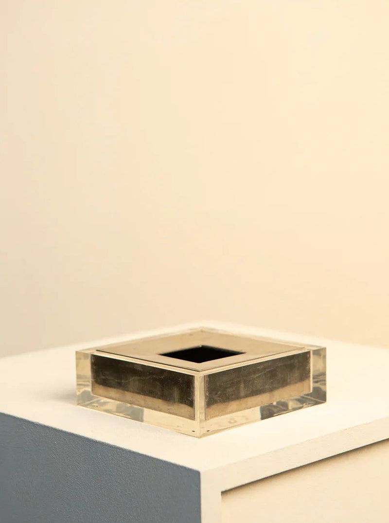A square-shaped Metal and Plexiglas Ashtray by Guzzini 70's from the Treaptyque brand features a metallic finish on a light-colored surface. This vintage-inspired piece, with its hollow central square and transparent outer layer, rests against a softly lit background, evoking a minimalist and clean aesthetic reminiscent of classic Italian design by Guzzini.