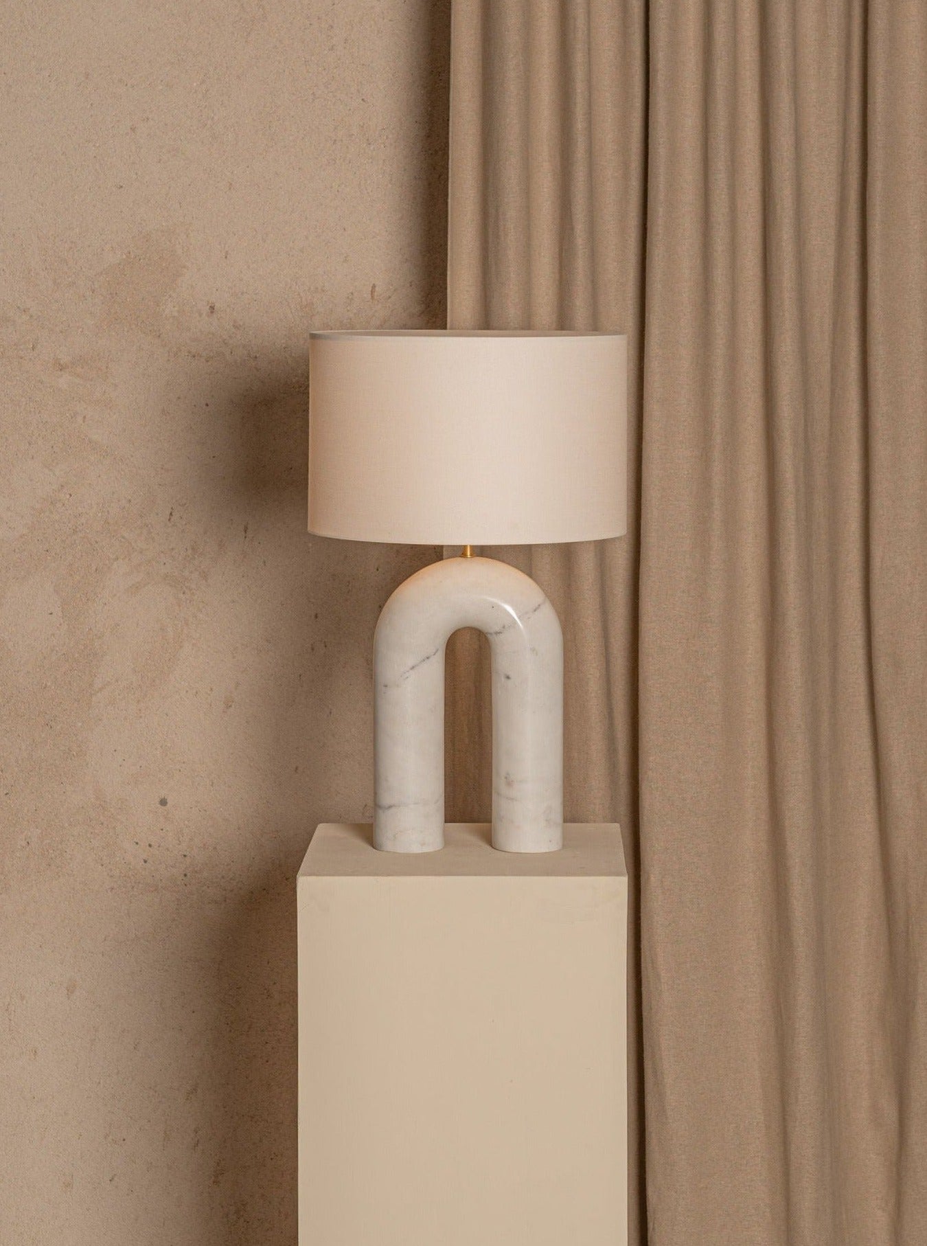 A modern Arko White Marble Lamp by Simone & Marcel with a curved base and a cotton lampshade stands on a beige pedestal against a textured, beige curtain backdrop.