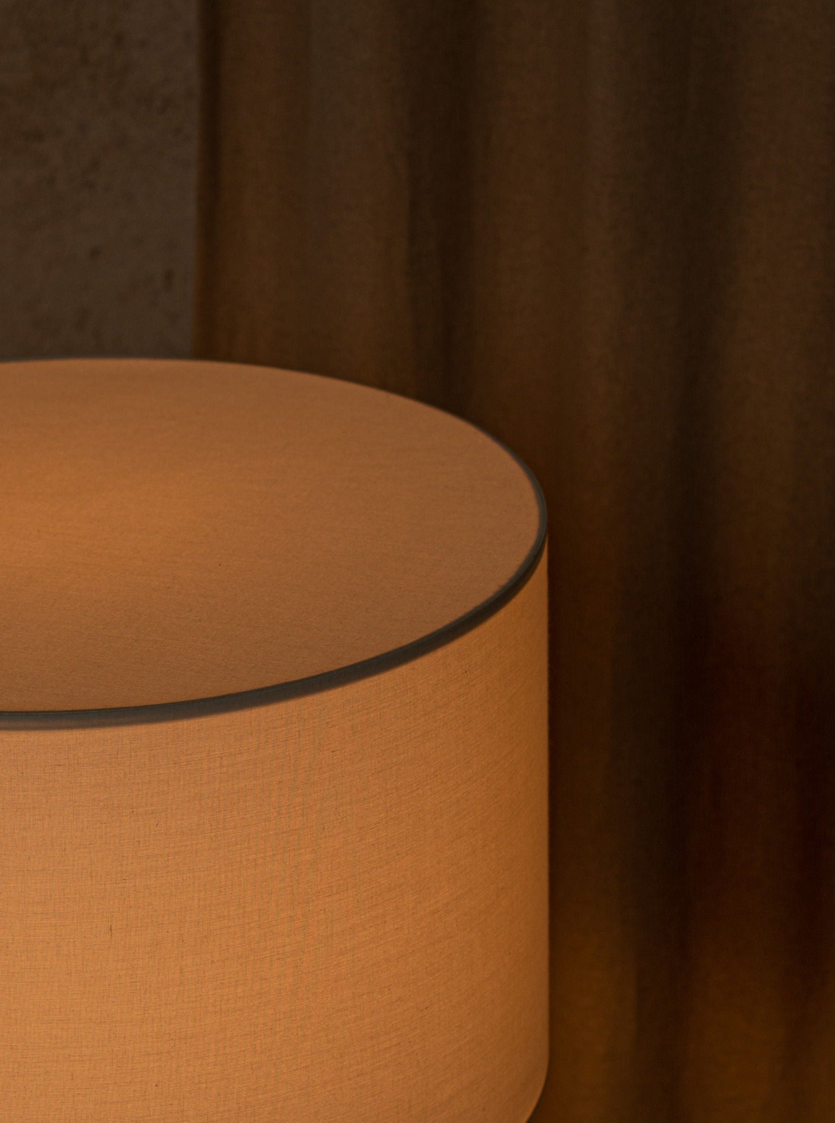 A close-up of a textured cylindrical Simone & Marcel Peona Black Marble table lamp against a curtain with warm, muted brown tones. The lighting casts soft shadows on the scene.