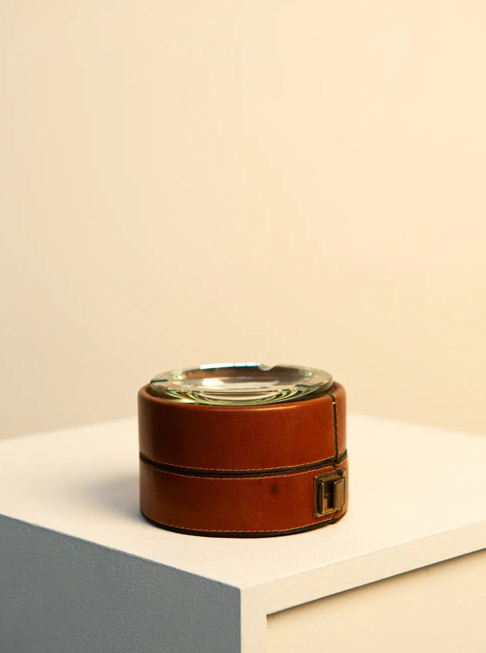 A round Italian Ashtray in brown leather and 60's crystal by Treaptyque rests on a beige and white pedestal. The soft lighting and neutral background give the scene a minimalist and elegant appearance, reminiscent of 60s craftsmanship.