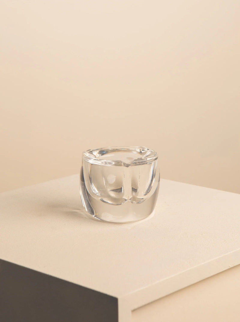 An elegant, clear Treaptyque crystal ashtray positioned on a gray surface, showcased against a light beige background. The rounded ashtray features thick walls and indentations for resting cigarettes.