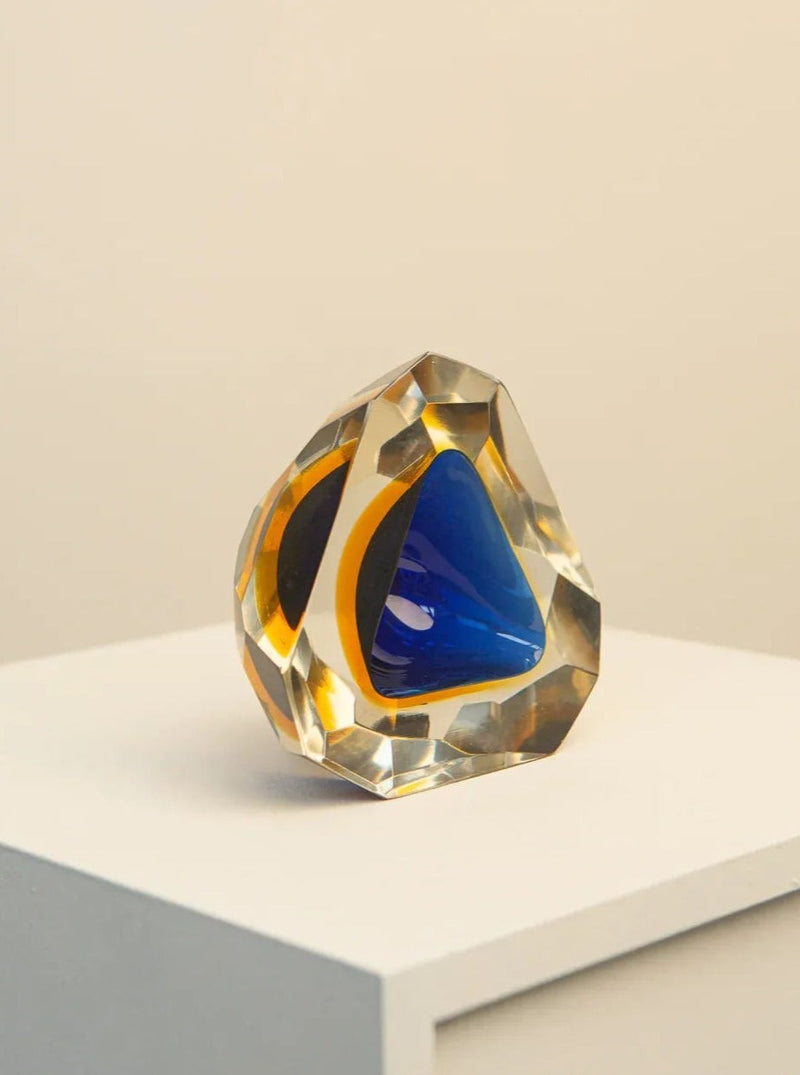 A geometric, multifaceted Treaptyque Multi-colour Ashtray in Murano Glass by Flavio Poli 60's with a blue triangular center is displayed on a white pedestal against a neutral background. Reminiscent of Flavio Poli's work, the outer layer is clear with golden-yellow accents, creating a prismatic effect.