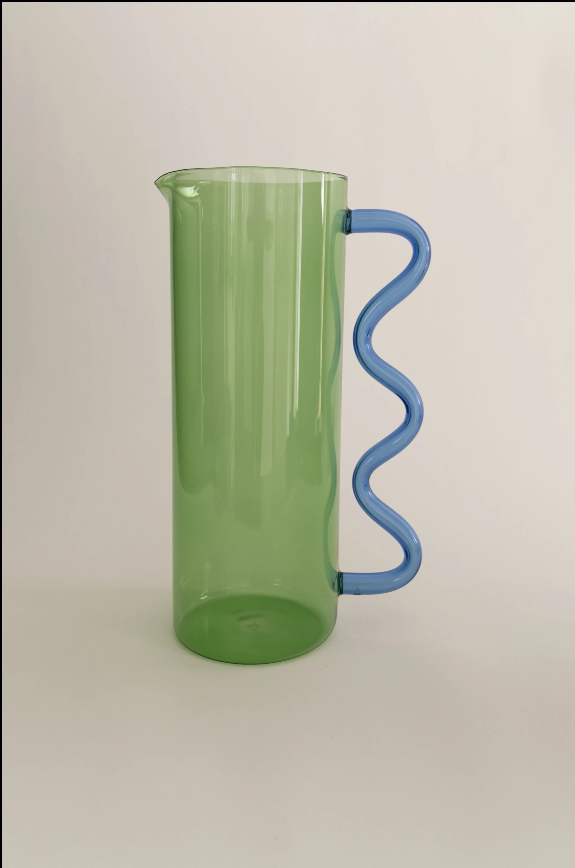 A versatile Sophie Lou Jacobsen Wave Pitcher Glass in green with a wavy blue handle, standing against a plain light background.