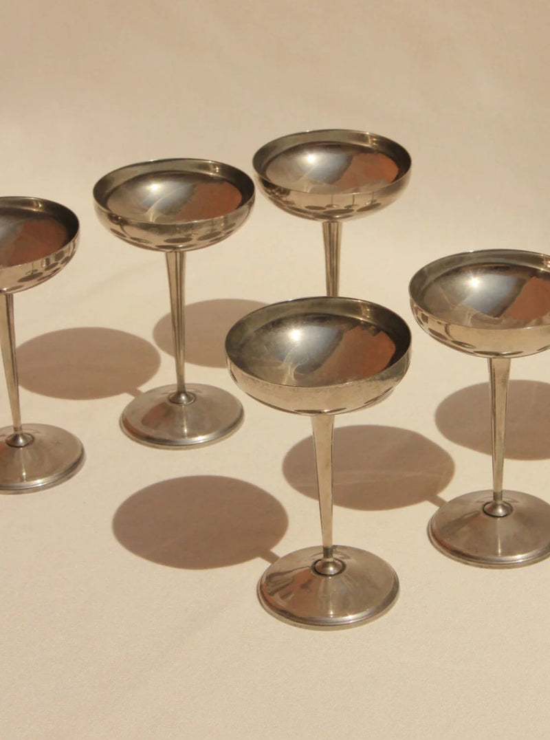 Five vintage Boga Avante Shop Alpaca Silver champagne coupes arranged on a sandy beige surface, showcasing their reflective bowls and elegant stems under soft lighting.