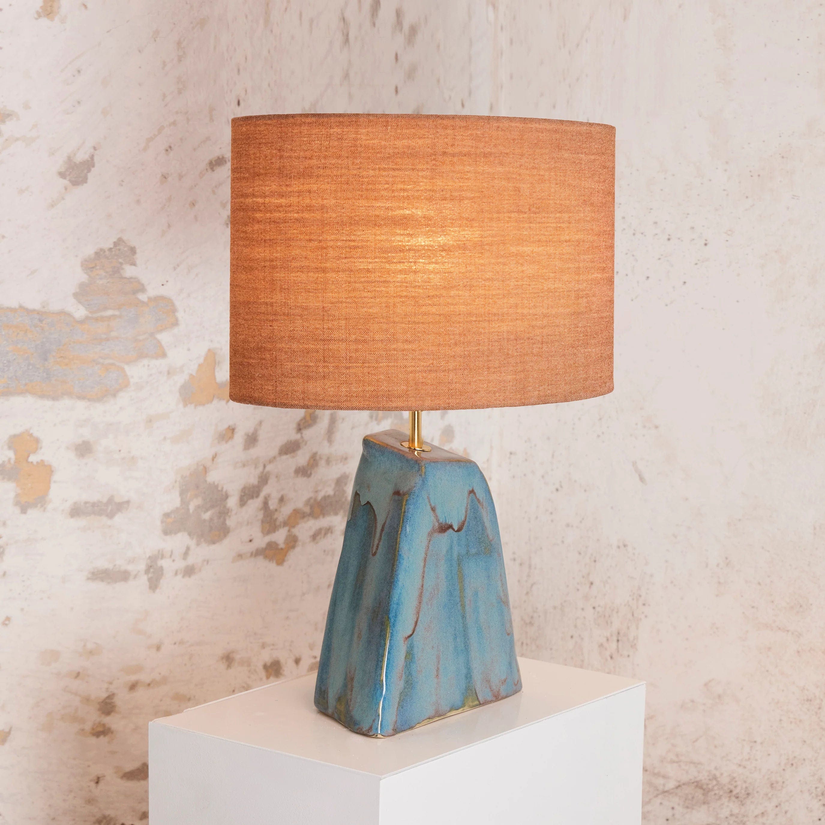 A Triangular Ceramic Lamp with unique blue and copper base by Project 213A and a cylindrical orange shade, illuminated and placed on a white pedestal against a textured wall.