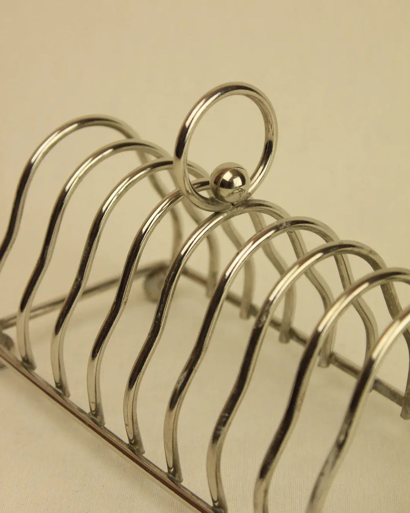 A close-up of a Boga Avante Shop Art Deco Toast Holder, featuring curved slots for bread and a central ring handle, displayed against a beige background.