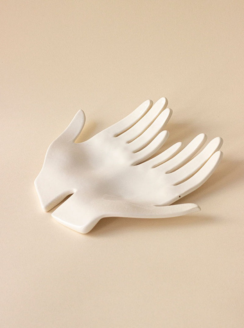 A white enamel Main Double Blanche dish shaped like a bird with outstretched wings against a plain beige background. The dish features slots that mimic the separation between the bird's feathers.