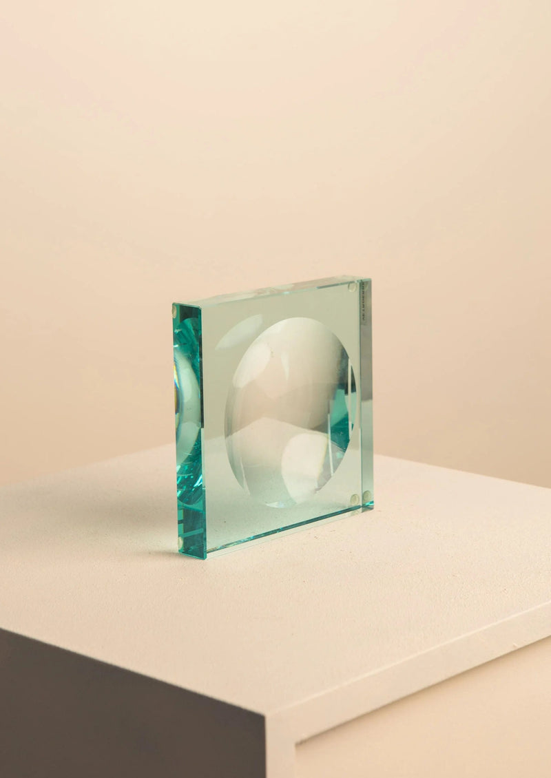 The Glass Vide Poche by Fontana Arte from Treaptyque is a handcrafted, transparent aqua-tinted glass cube with a circular indentation, elegantly resting on a beige pedestal, embodying minimalist beauty against a soft, neutral backdrop.