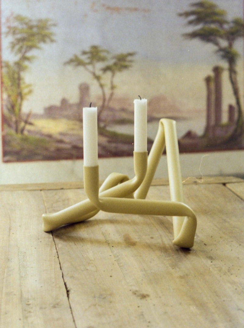 A modern, abstract Bucatini Candle Holder (Beige) from AOT STUDIO with two white candles stands on a wooden surface. The background features a pastoral landscape painting with trees, water, and ancient ruins, highlighting the piece's exquisite Danish craftsmanship.