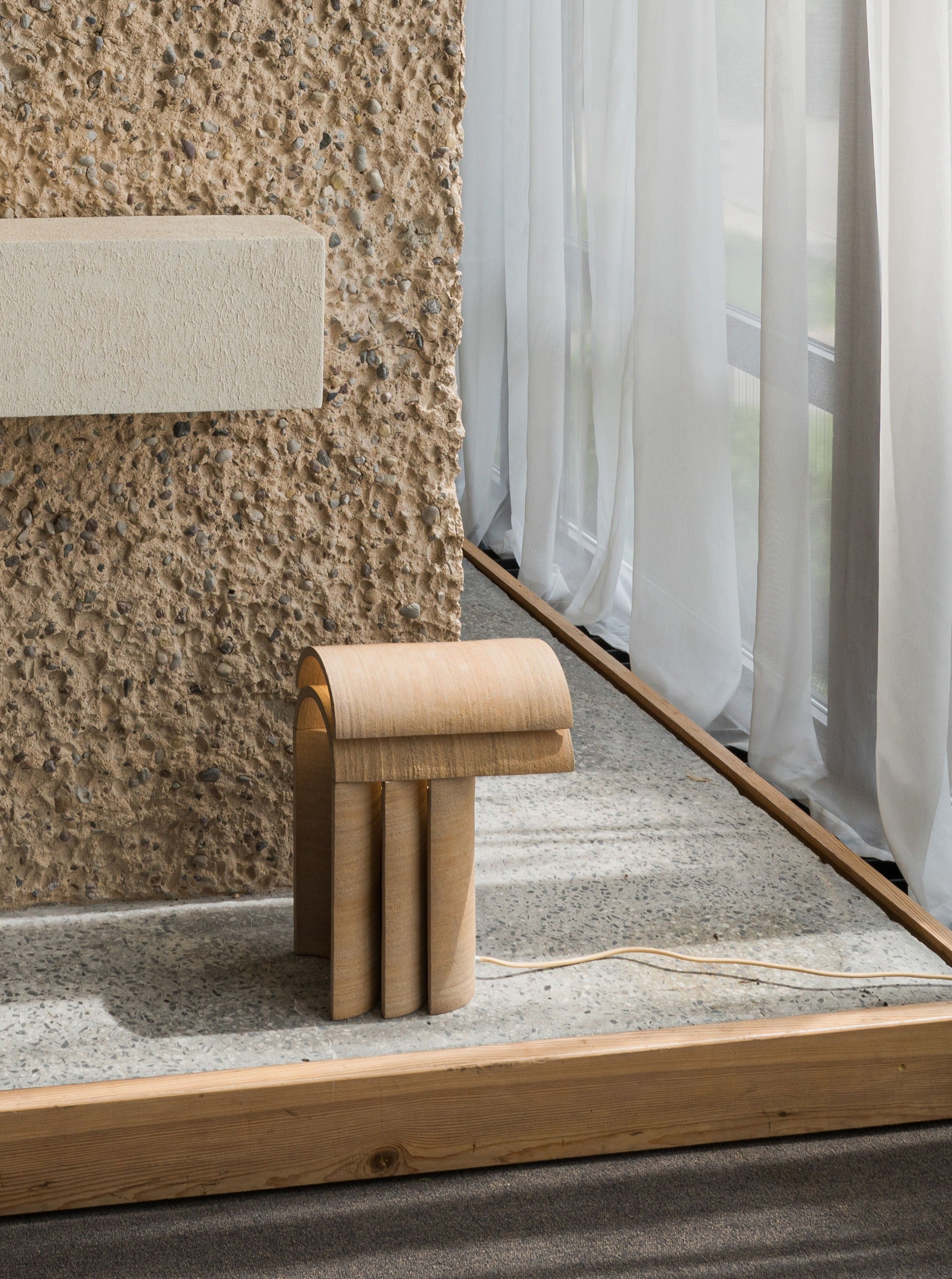 A minimalist wooden chair with a curved backrest and cylindrical legs stands beside a window with sheer curtains, against a textured stone wall and sculptural lighting highlighting the terrazzo floor by Evelina Kudabaite's "Layers of..." Light I.