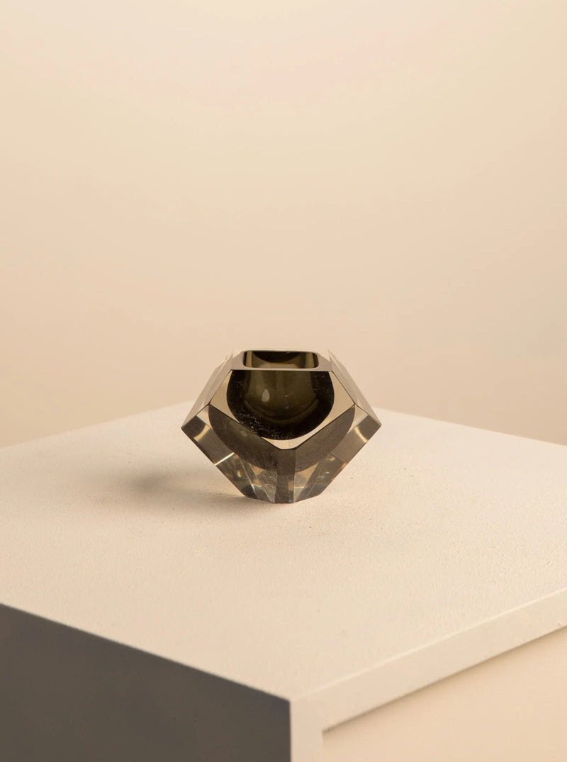 A black "Diamant" ashtray in Murano glass by Flavio Poli for Seguso from the 60s, offered by Treaptyque, sits on a white pedestal. Its small geometric design features sharp, angled facets and a transparent base with a slight grey tint, casting subtle shadows on the pedestal and background to create a minimalist aesthetic.