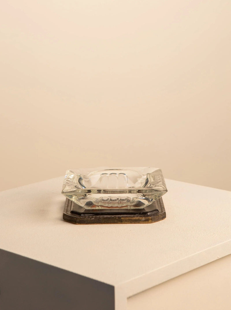 The Treaptyque Saint-Louis Crystal and Leather Ashtray, with its sculptural square design, rests on a white surface. The soft beige background creates a luxurious and minimalist scene perfect for sophisticated interiors.