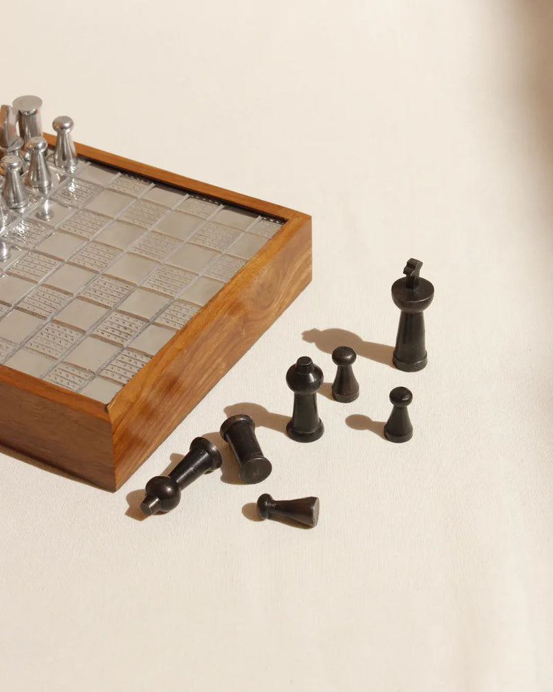 The Boga Avante Shop’s Wood and Metal Chessboard boasts a collectible wooden design with a silver metallic grid, elegantly displayed on a light surface. Black chess pieces such as pawns and the queen are artfully scattered, with side lighting enhancing its luxurious interior appeal.