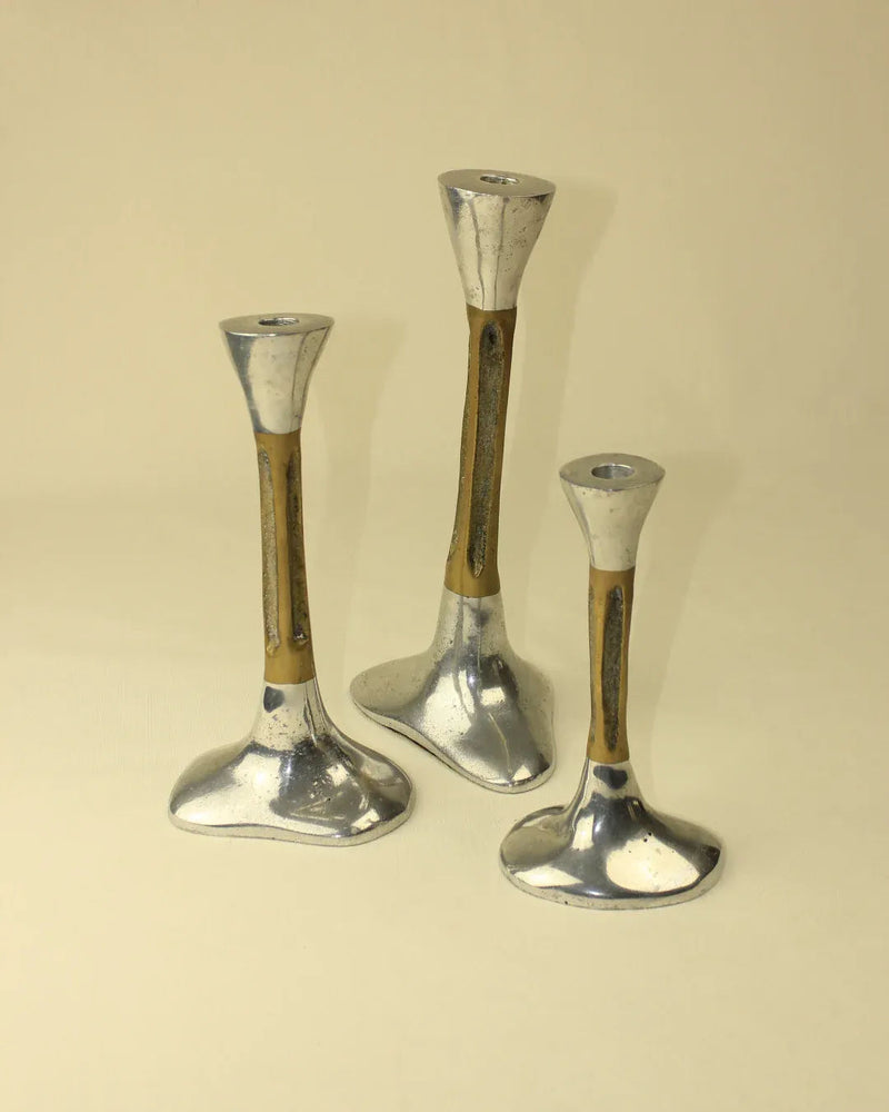 The Trio of Brutalist Candleholders from Boga Avante Shop are arranged on a beige surface. Each candleholder showcases a flared, rounded base, a slim, slightly tapered middle section, and a wide, shallow holder at the top. The metal boasts a subtly distressed, vintage look that harkens back to 1980s design. Ideal for adding character to your home decor.