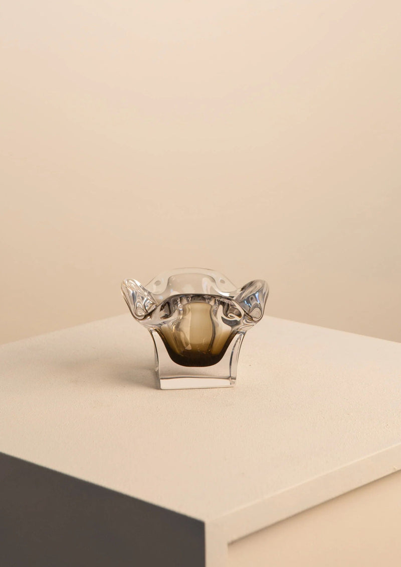 A small, transparent handcrafted Treaptyque Murano Flower Vide Poche with a unique wavy rim and thick base, displayed on a light grey podium against a beige background.