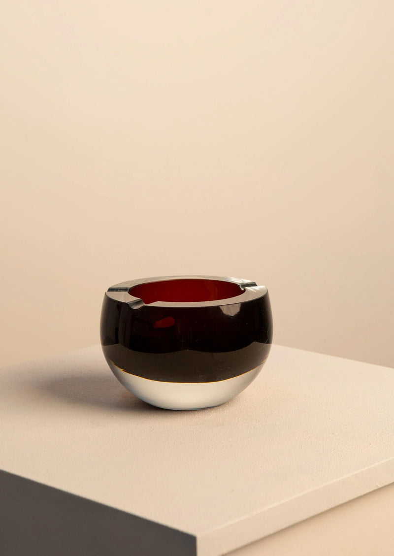 The Large Murano Glass Ashtray by Treaptyque rests on a light surface. It boasts a handcrafted, sculptural design with a reflective black exterior, contrasting red interior, and elegant rim notches. The minimalist aesthetic is complemented by the plain muted beige background.