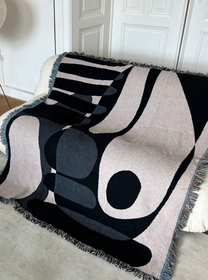 A modern Rokko Jacquard Throw with abstract geometric designs in black, gray, and blush colors, draped over a white door, featuring a textured fringe border and crafted as a luxury throw by 2222Studio.