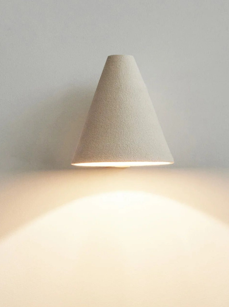 A minimalist cone-shaped wall sconce emitting a soft glow along its bottom edge, casting a warm circular reflection on a neutral wall and surface., Mariza Galani's Esoteric Wall Lamp.