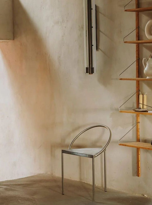 A minimalist room with a simple, modern Triangolo Chair | Stainless Steel by FRAMA featuring an arched backrest. The wall behind the chair is plain, and to the right, there's a wooden shelving unit with various ceramic vases and books. Sunlight softly illuminates the space, enhancing its industrial aesthetic.